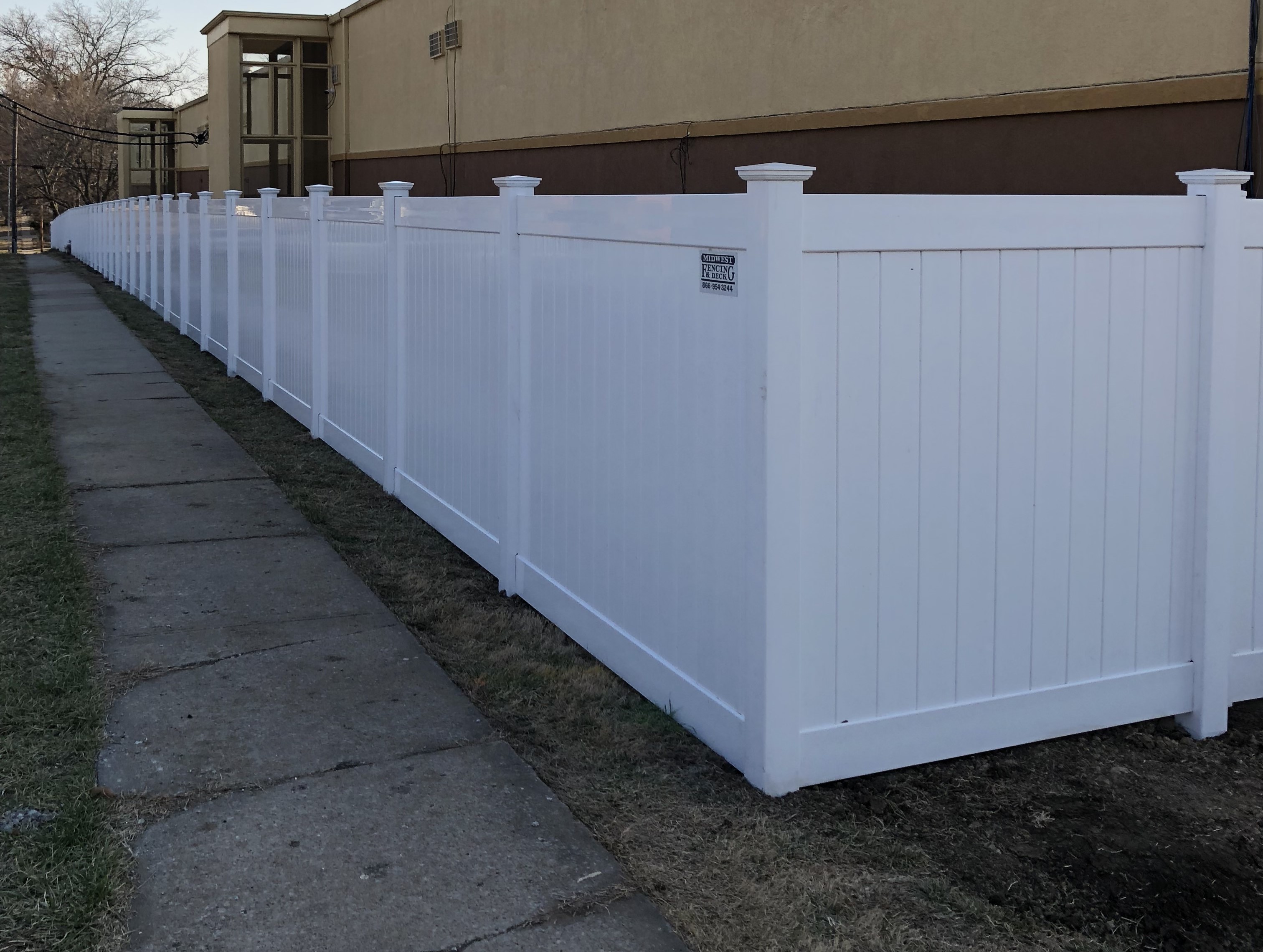tall white vinyl fence with one bush in front along a road